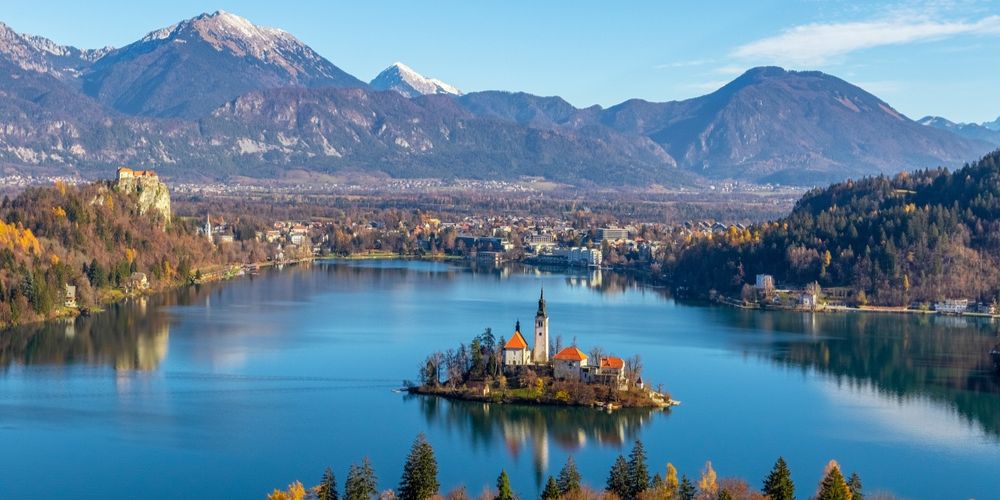 The serene Lake Bled in Slovenia at sunrise, showing the iconic island church reflected in still, emerald waters with misty mountains in the background.